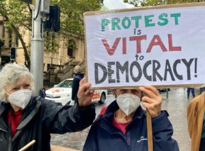 Two elderly activists hold a sign reading: Protest is vital to democracy