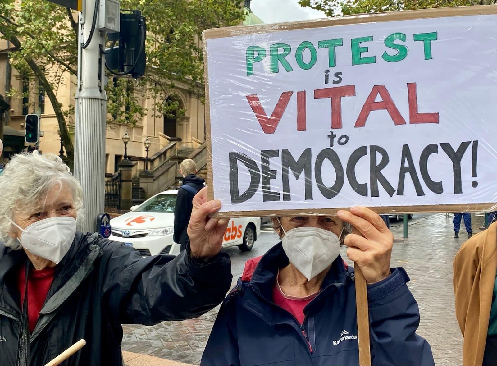 Two elderly activists hold a sign reading: Protest is vital to democracy!