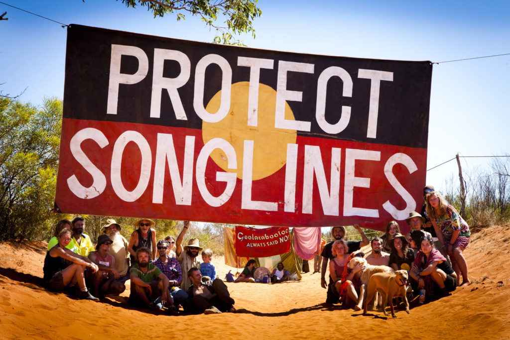 A huge banner reading Protect Songlines with activists seated below in the red dirt of the Kimberley