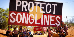 Banner reading Protect Songlines with activists seated beneath in the red dirt of the Kimberley