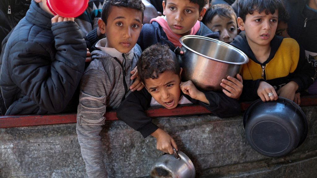 A crowd of children are squashed together with pots in their hands looking distressed. Caption from article: Palestinian children wait to receive food cooked by a charity in Rafah, southern Gaza. Pic: Reuters 